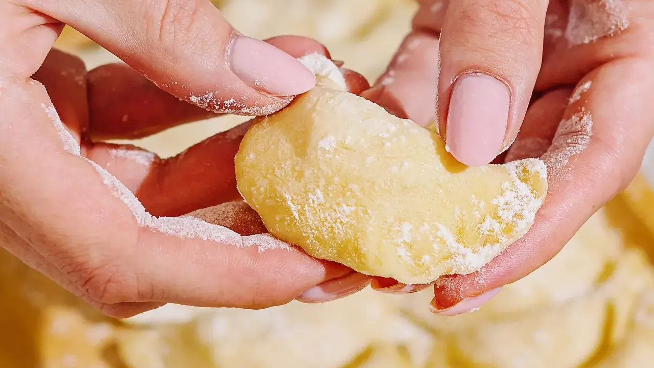 Dłonie formują ciasto na pierogi, posypane mąką. W tle widać gotowe pierogi.