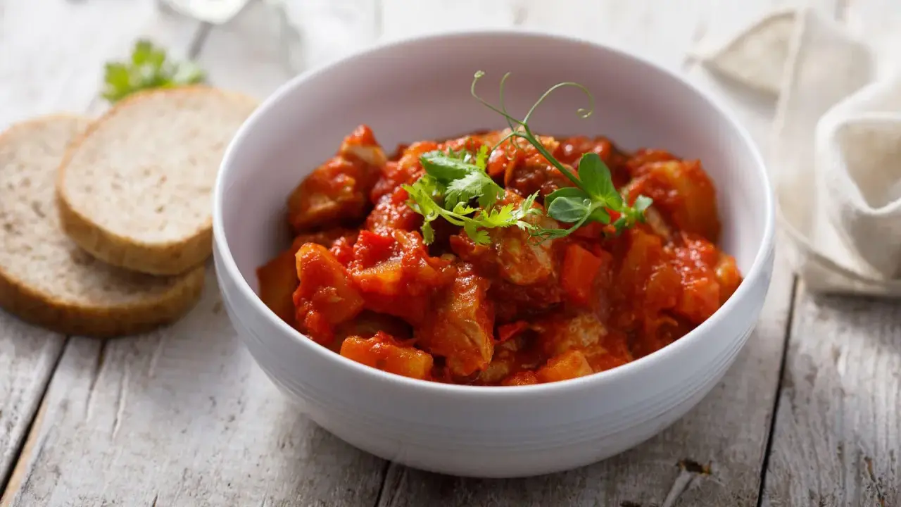Aromaticzny indyk w sosie curry, podany w białej misce, z dodatkiem świeżych ziół i kromek chleba.