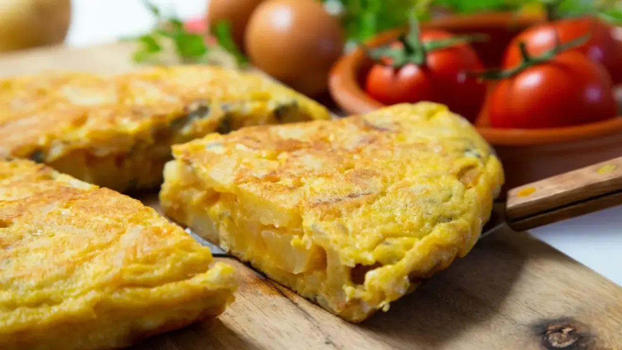 Pyszna tortilla de patatas, pokrojona na kawałki, leży na desce. W tle jajka, pomidory i natka pietruszki.