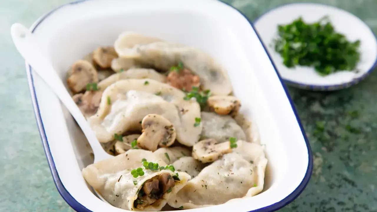 Pyszne pierogi z pieczarkami, podane w białej misce z niebieską obwódką, ozdobione natką pietruszki. Obok talerzyk z posiekanymi ziołami.