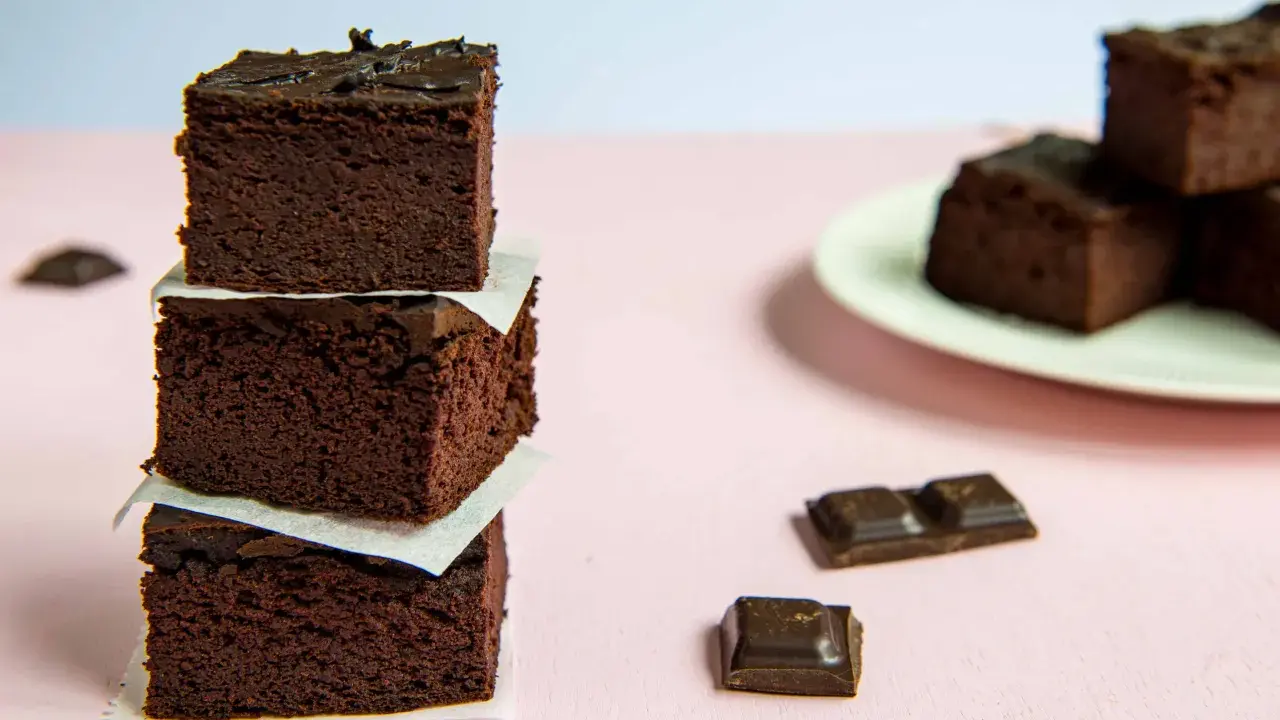 Ciemne, wilgotne brownie z fasoli ułożone w wieżę, obok kawałki czekolady i talerz z kolejnymi ciastkami.
