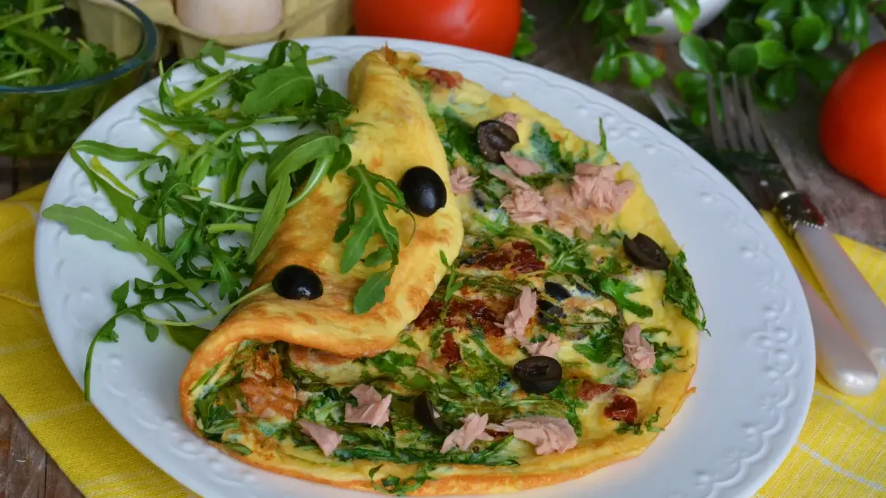 Pyszny omlet z tuńczykiem, rukolą i oliwkami, podany na białym talerzu z żółtym obrusem.