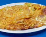 Złocisty omlet na słono z kawałkami kiełbasy i zielonych warzyw, podany na białym talerzu.
