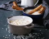 Gotowy do podania, kremowy ryż na mleku w ceramicznej miseczce, obok miseczki z surowym ryżem i laskami cynamonu.