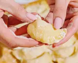Dłonie formują ciasto na pierogi, posypane mąką. W tle widać gotowe pierogi.