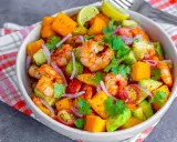 Kolorowa sałatka z krewetkami, awokado, mango, pomidorkami i czerwoną cebulą, udekorowana kolendrą i ćwiartkami limonki.