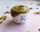 Gładka, zielona pasta pistacjowa w białej filiżance z niebieskimi kropkami. Wokół rozsypane pistacje. Idealna do domowego wypieku, jak zrobić pastę pistacjową.