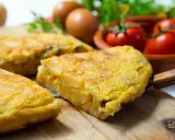 Pyszna tortilla de patatas, pokrojona na kawałki, leży na desce. W tle jajka, pomidory i natka pietruszki.