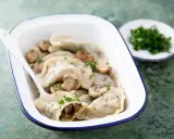 Pyszne pierogi z pieczarkami, podane w białej misce z niebieską obwódką, ozdobione natką pietruszki. Obok talerzyk z posiekanymi ziołami.