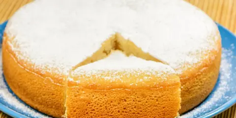 Puszyste ciasto bez mleka, posypane cukrem pudrem, z wyciętym kawałkiem, prezentuje się apetycznie na niebieskim talerzu.