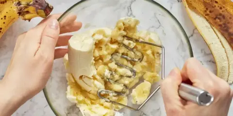 Ręce ugniatają banany w misce, przygotowując pyszny deser z bananów. Obok leżą dojrzałe banany.