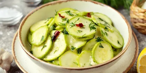 Pyszna surówka z cukinii z koperkiem i chili, podana w misce. Obok czosnek i cytryna.