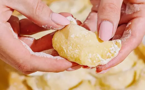 Dłonie formują ciasto na pierogi, posypane mąką. W tle widać gotowe pierogi.