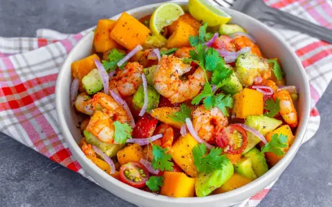 Kolorowa sałatka z krewetkami, awokado, mango, pomidorkami i czerwoną cebulą, udekorowana kolendrą i ćwiartkami limonki.