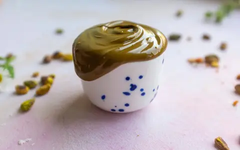 Gładka, zielona pasta pistacjowa w białej filiżance z niebieskimi kropkami. Wokół rozsypane pistacje. Idealna do domowego wypieku, jak zrobić pastę pistacjową.