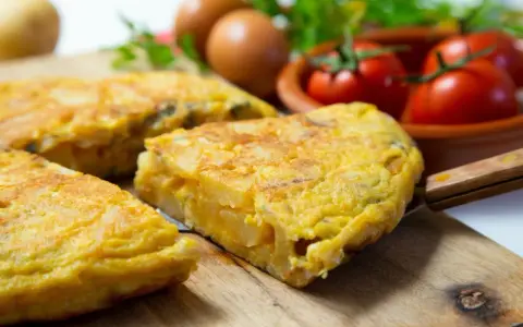 Pyszna tortilla de patatas, pokrojona na kawałki, leży na desce. W tle jajka, pomidory i natka pietruszki.
