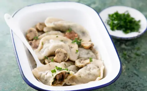 Pyszne pierogi z pieczarkami, podane w białej misce z niebieską obwódką, ozdobione natką pietruszki. Obok talerzyk z posiekanymi ziołami.