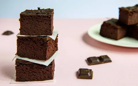 Ciemne, wilgotne brownie z fasoli ułożone w wieżę, obok kawałki czekolady i talerz z kolejnymi ciastkami.
