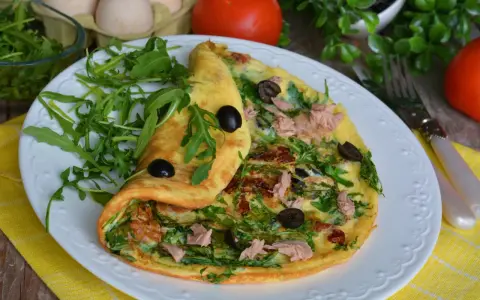 Pyszny omlet z tuńczykiem, rukolą i oliwkami, podany na białym talerzu z żółtym obrusem.