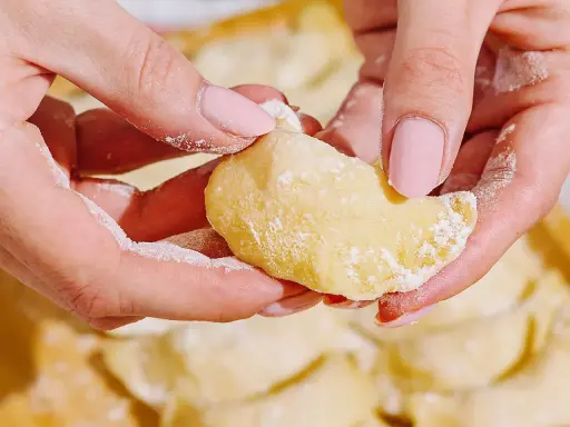 Dłonie formują ciasto na pierogi, posypane mąką. W tle widać gotowe pierogi.