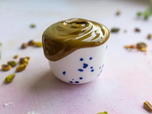 Gładka, zielona pasta pistacjowa w białej filiżance z niebieskimi kropkami. Wokół rozsypane pistacje. Idealna do domowego wypieku, jak zrobić pastę pistacjową.