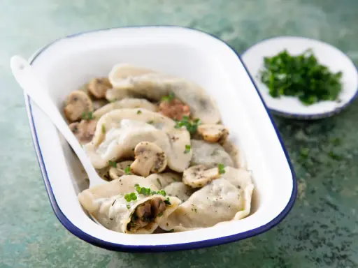 Pyszne pierogi z pieczarkami, podane w białej misce z niebieską obwódką, ozdobione natką pietruszki. Obok talerzyk z posiekanymi ziołami.