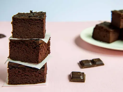 Ciemne, wilgotne brownie z fasoli ułożone w wieżę, obok kawałki czekolady i talerz z kolejnymi ciastkami.