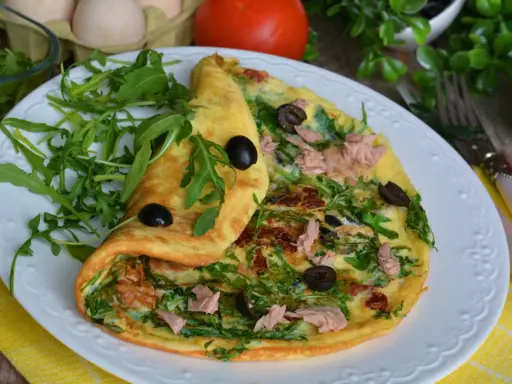 Pyszny omlet z tuńczykiem, rukolą i oliwkami, podany na białym talerzu z żółtym obrusem.
