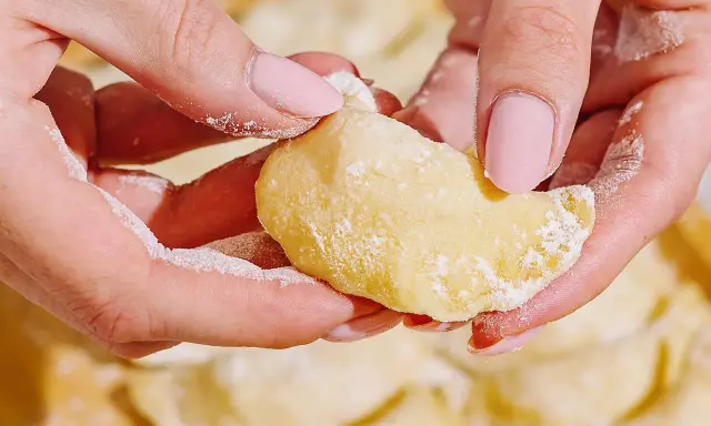 Dłonie formują ciasto na pierogi, posypane mąką. W tle widać gotowe pierogi.
