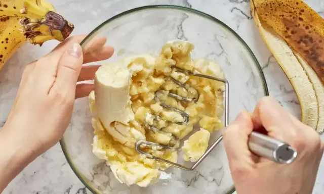 Ręce ugniatają banany w misce, przygotowując pyszny deser z bananów. Obok leżą dojrzałe banany.