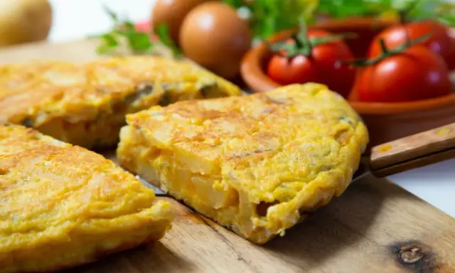 Pyszna tortilla de patatas, pokrojona na kawałki, leży na desce. W tle jajka, pomidory i natka pietruszki.