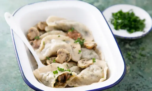 Pyszne pierogi z pieczarkami, podane w białej misce z niebieską obwódką, ozdobione natką pietruszki. Obok talerzyk z posiekanymi ziołami.