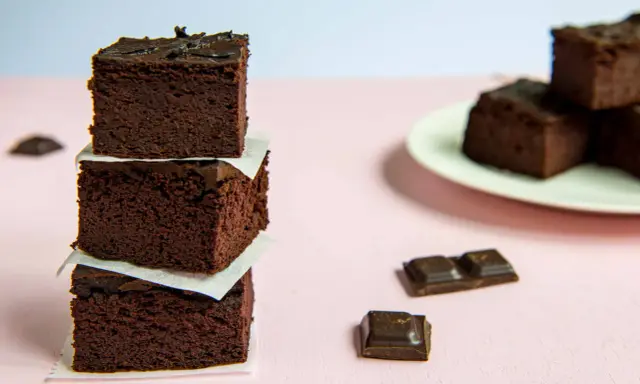 Ciemne, wilgotne brownie z fasoli ułożone w wieżę, obok kawałki czekolady i talerz z kolejnymi ciastkami.