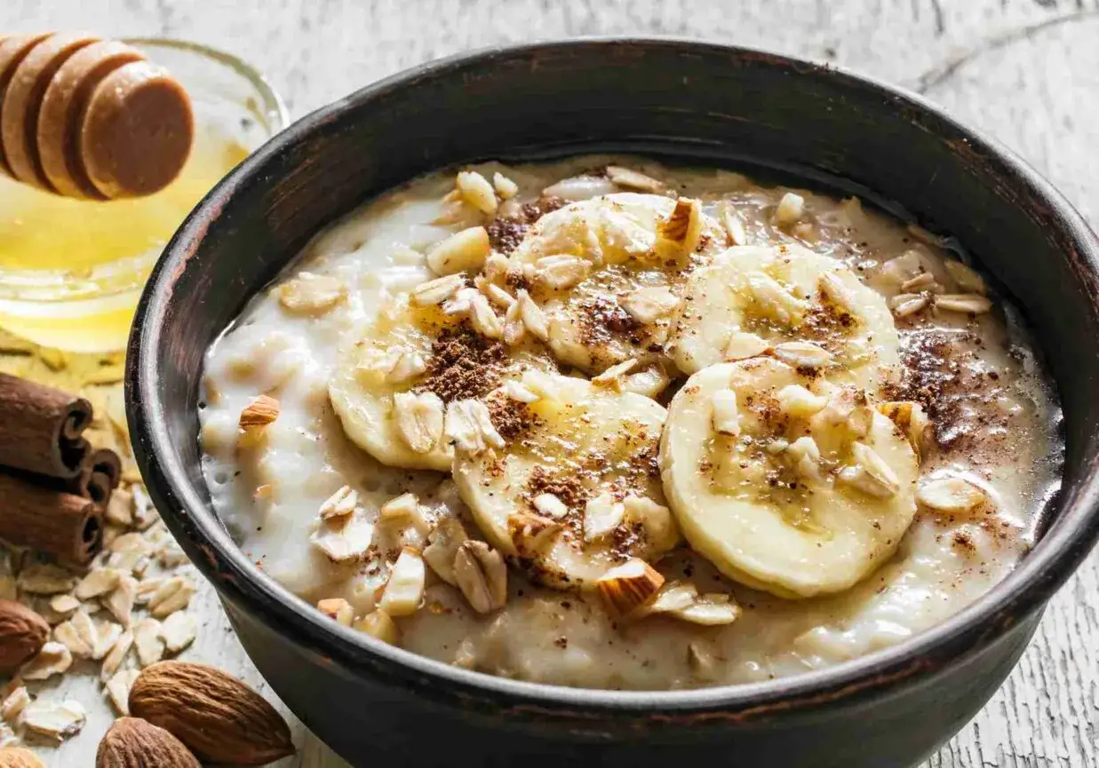 Pyszna owsianka z bananem, posypana płatkami migdał&oacute;w i cynamonem. Obok mi&oacute;d i laskowe orzechy.