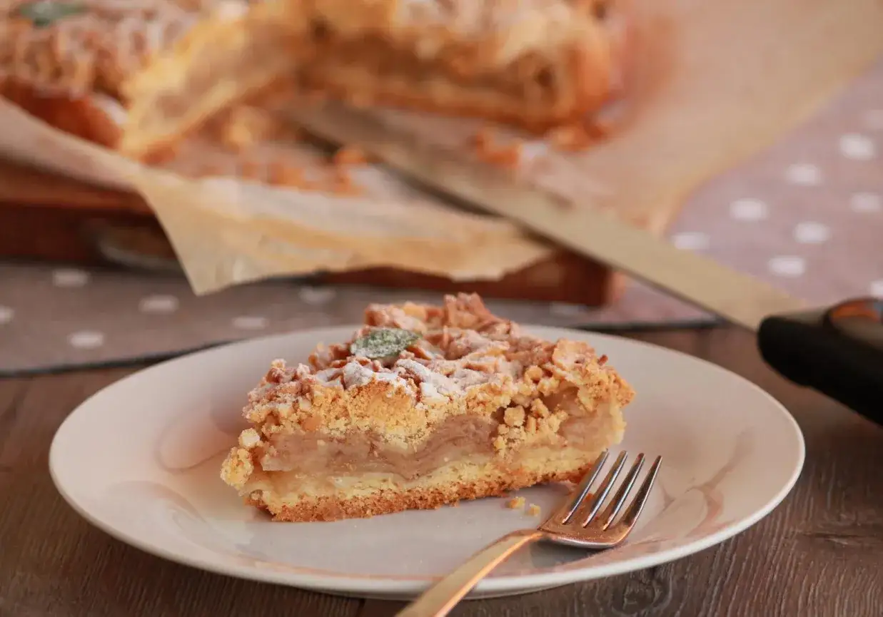 Pyszna tarta z jabłkami, posypana cukrem pudrem i listkiem mięty, czeka na talerzyku. Obok leży widelec, gotowy do degustacji.