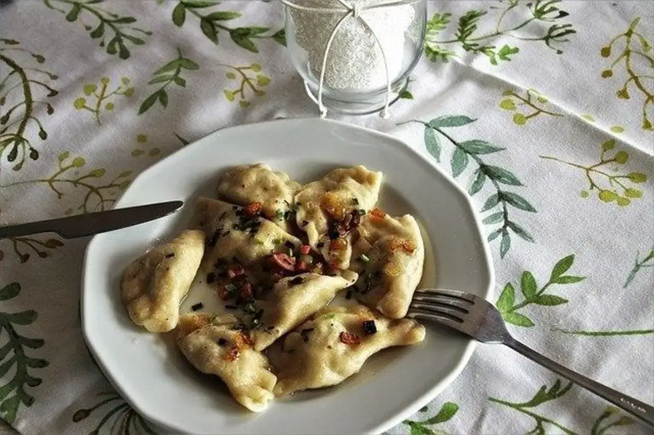 Pyszne pierogi z pieczarkami, polane skwarkami i posypane szczypiorkiem, czekają na talerzu. Obok n&oacute;ż i widelec.