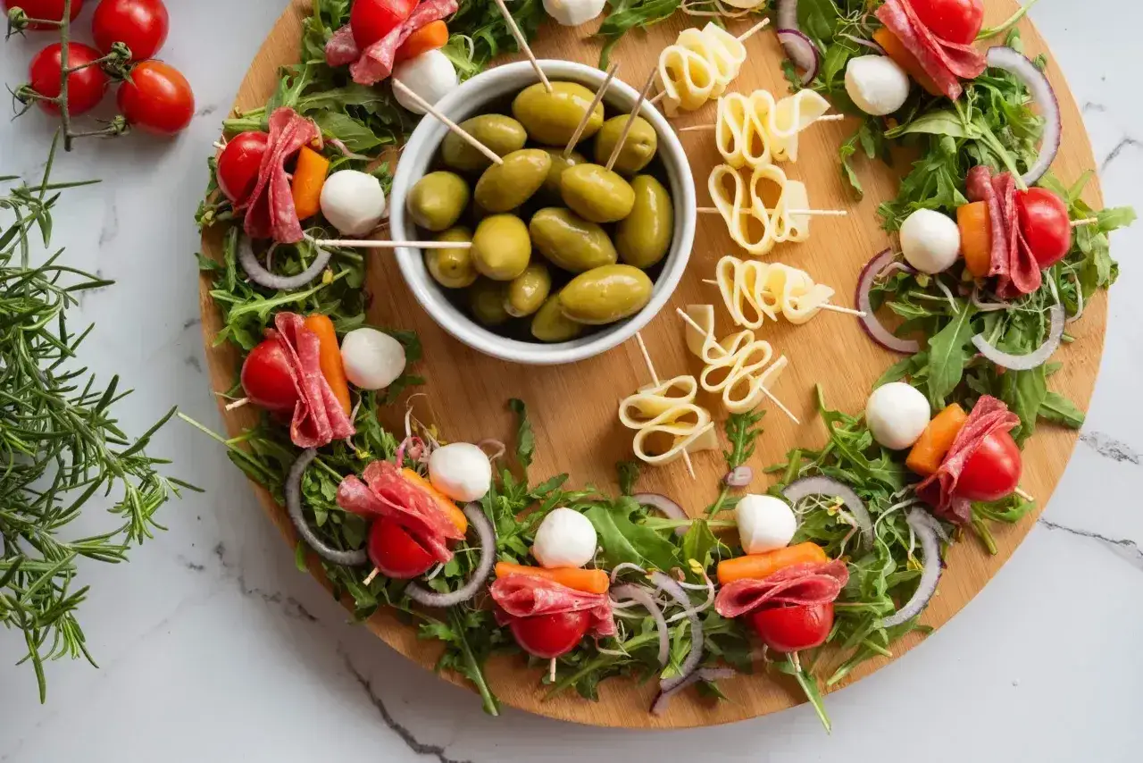 Kolorowa sałatka na imprezę: szaszłyki z pomidorkami, mozzarellą, salami i marchewką, oliwki i ser na desce.