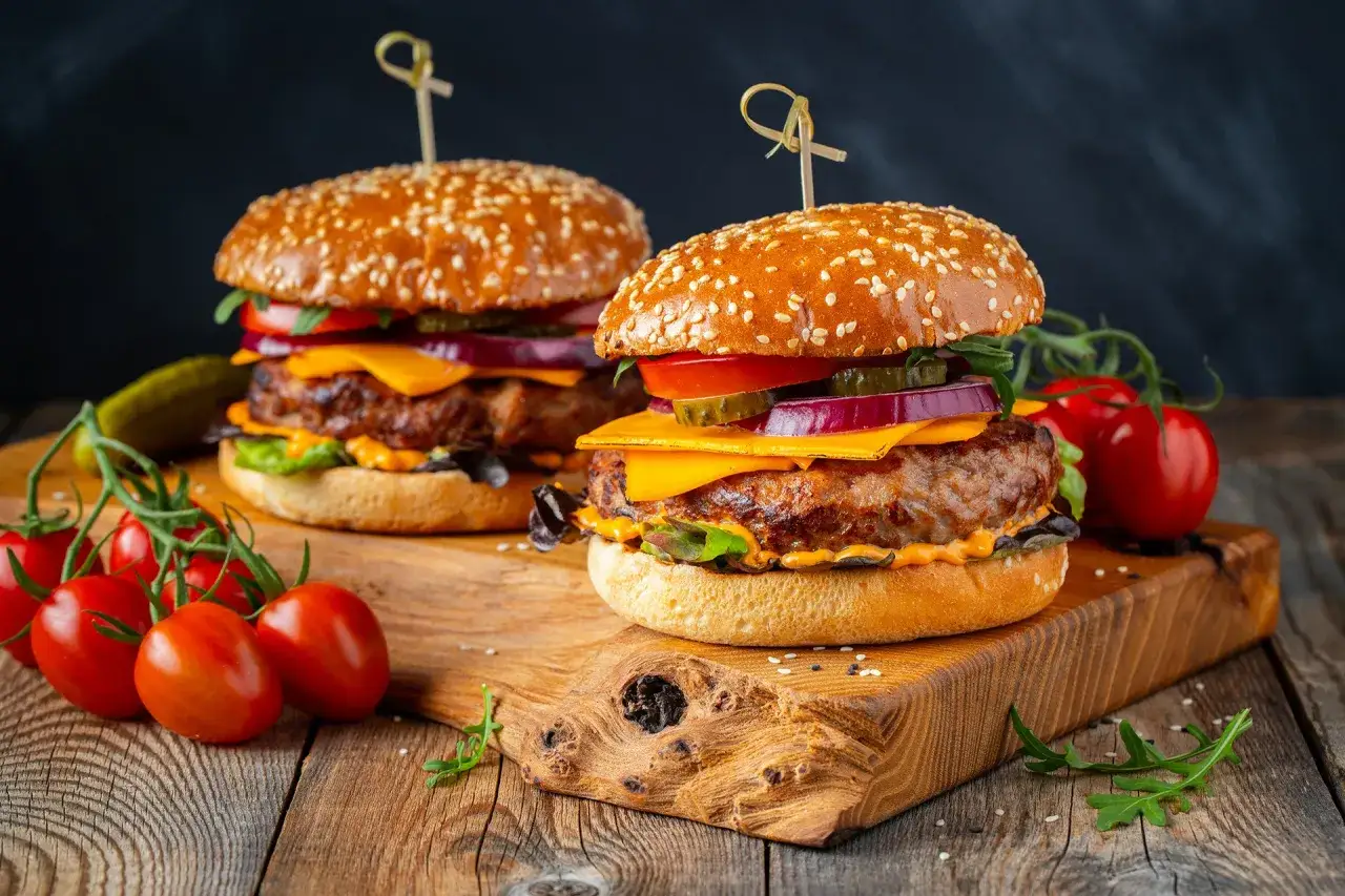 Dwa soczyste burgery z idealnie wysmażonym mięsem na burgery, serem, warzywami i sezamową bułką, podane z pomidorkami na drewnianej desce.