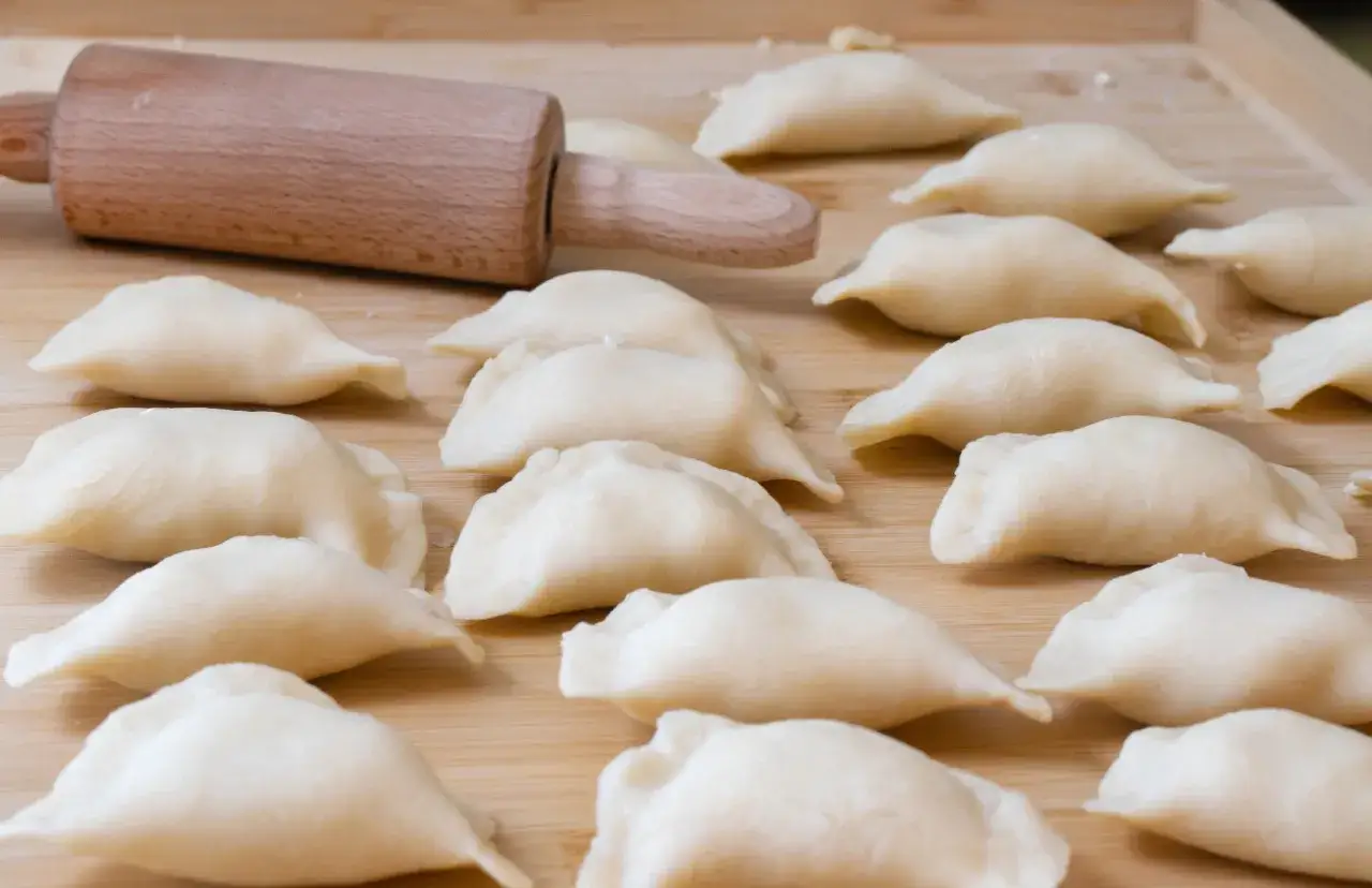Gotowe do lepienia pierogi, ułożone rzędami na drewnianej desce, obok wałka.